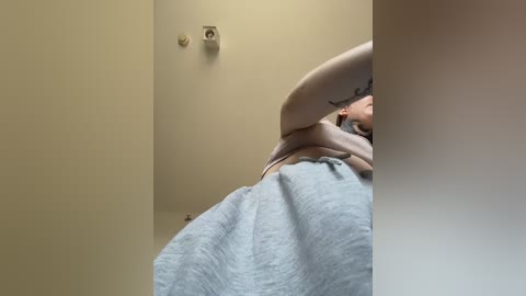 Media: A video of a person with light skin, wearing a gray sweater, captured from a low angle, showing their upper torso and arm, partially obscured by a wall.