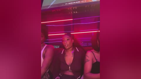 Video of a Black woman with medium skin tone, medium-length braids, and tattoos, wearing a dark zip-up jacket and jeans, sitting in a dimly-lit nightclub. Background features neon lights and a partially visible sign.