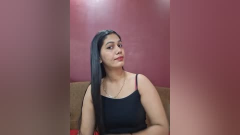 Media: Video of a young woman with long, dark hair, wearing a black spaghetti-strap top, sitting against a pink wall in a dimly lit room.