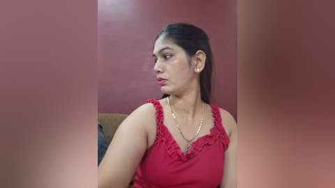 Video of a young South Asian woman with medium skin tone and straight black hair, wearing a red sleeveless top with ruffled neckline, seated in a room with red walls, looking contemplative.