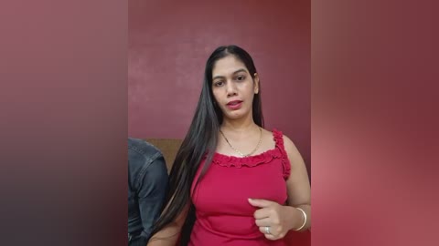 Video of a smiling, medium-skinned woman with long black hair, wearing a red sleeveless top, seated next to a man in jeans on a brown couch against a red wall.