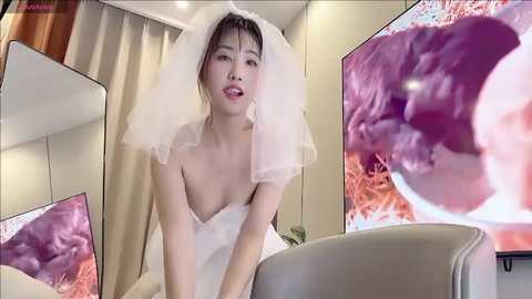 Media: Video of an Asian woman in a white wedding dress with a veil, smiling in a modern, minimalist room with beige walls, featuring a large TV screen displaying an abstract, colorful, swirling pattern.