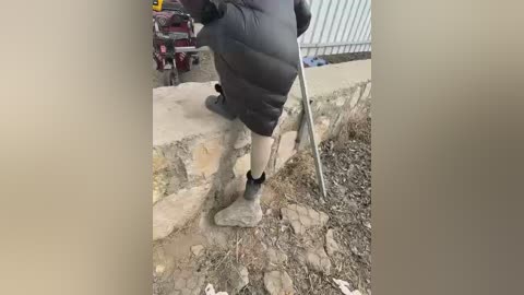 Video of a person in a black puffer jacket, beige pants, and black shoes, using a shovel to clean a stone wall. Background shows a red cart and industrial structures.