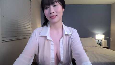 Media: Video of an Asian woman with straight black hair, fair skin, and a slender physique, wearing a white button-up shirt partially unbuttoned, revealing cleavage, in a dimly lit bedroom with a bed, nightstand, and window.