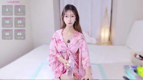 Video of a young Asian woman with long brown hair, wearing a pink floral kimono, sitting on a bed in a minimalist, well-lit bedroom, holding a water bottle.