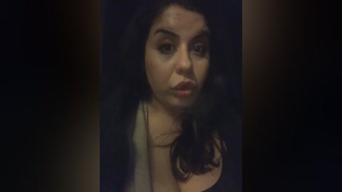 A dimly lit video of a woman with dark hair and fair skin, wearing a black top, looking solemn against a dark background.