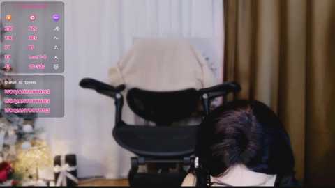 Video of a woman in a black chair with a beige blanket, in a room with white curtains and a toy display.