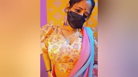 Video of a South Asian woman in traditional yellow floral blouse, black face mask, and pink sari with blue border, against a yellow patterned wall, wearing red earrings.