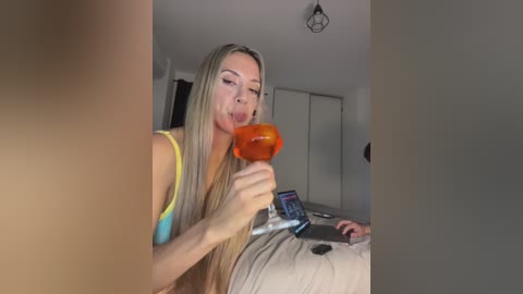 Video of a blonde woman in a yellow tank top drinking a bright orange beverage, seated on a bed with a phone on her lap, in a modern, minimally decorated bedroom.