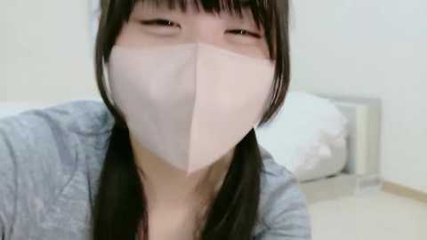 A video of an East Asian woman with long black hair, wearing a light blue shirt and a white face mask, smiling. The background shows a white wall and a bed with white sheets.