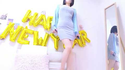 Video of a slender, light-skinned woman with straight black hair, wearing a light blue dress and cardigan, standing in a bright, minimalist room with gold \"Happy New Year\" letters and a mirror.