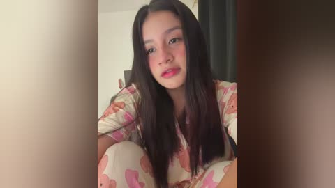 Media: Video of a young East Asian woman with long black hair, wearing a floral-patterned shirt, sitting on a bed with a blurred background.