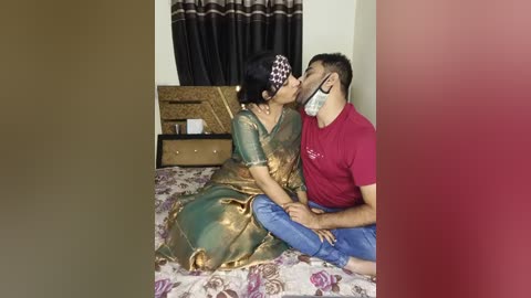 Video of an Indian couple kissing passionately on a bed, the woman in a gold sari, the man in a red t-shirt, both wearing masks, in a modest bedroom with floral bedding and a wooden dresser.