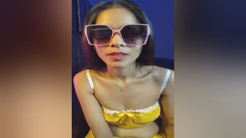 Video of a young Asian woman with long hair, wearing oversized pink sunglasses, yellow top, and sitting against a blue backdrop.