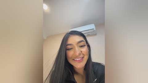 Video of a smiling Latina woman with long, straight black hair, wearing a black jacket, standing in a narrow, beige-walled hallway with an air conditioner on the wall above her.