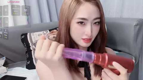 Media: Video of a young Asian woman with light skin and long brown hair, using a purple dildo on a grey couch with black and white pillows.