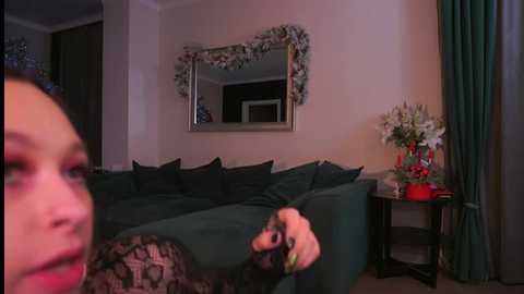 Video of a cozy living room with a woman holding a controller, a dark green sectional sofa, a large mirror with garlands, a floral arrangement, and teal curtains.
