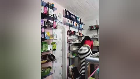A video of a cluttered, small kitchen with a person in a red and black striped shirt rummaging through the fridge. Shelves are packed with food items and bottles, and the white walls are plain.