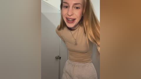 A video of a smiling young woman with straight blonde hair, wearing a beige long-sleeve top and light gray sweatpants, standing in a white doorway.