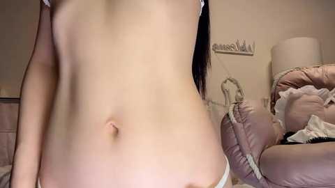 A video of a slender, topless Asian woman with light skin, wearing white panties, standing in a softly lit, pink-themed bedroom with a bed, lamp, and hanging clothes.