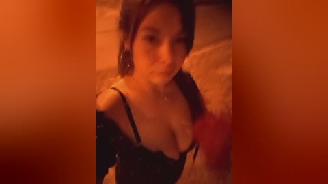 A dimly-lit video shows a woman with long, dark hair and fair skin, wearing a black off-shoulder dress, her cleavage partially exposed. She appears to be in a cozy, dimly-lit room with warm, orange lighting.