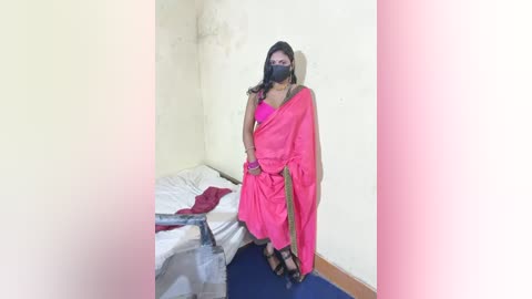 A video of a woman with medium brown skin, wearing a pink saree, black mask, and black high-heeled sandals, standing in a sparsely furnished room with white walls and a blue floor.