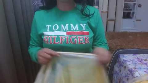 Video of a woman in a green long-sleeve shirt with \"TOMMY HILFIGER\" logo, holding a yellow and white striped fabric, standing indoors with a floral-patterned bed in the background.