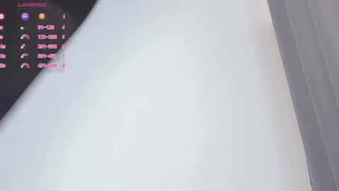 Video of a light gray wall with a black curtain partially covering the left side. A digital display on the left shows streaming information with pink text. The room appears minimalistic and modern.