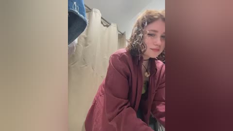 Video of a curly-haired, fair-skinned woman in a maroon robe, looking down in a beige-tiled bathroom with a blue towel and shower curtain.