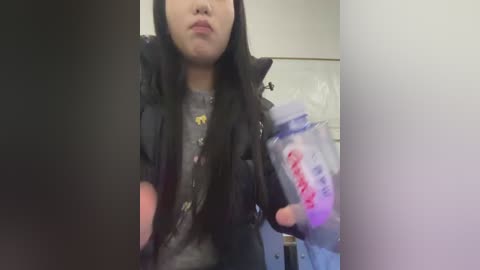 Video of a woman with long black hair, wearing a gray graphic tee and a black jacket, holding a bottle labeled \"Aptamil\" in a dimly lit bathroom.