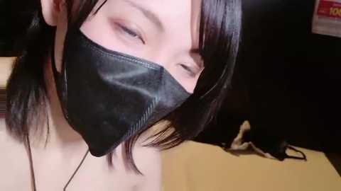 Video of a young woman with pale skin, wearing a black leather face mask and dark hair, in a dimly lit room with a black background.
