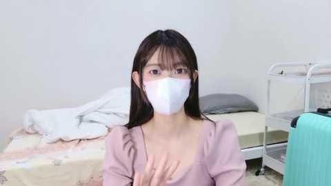 A video of an East Asian woman with long black hair, wearing a white surgical mask, pink blouse, and standing in a minimalistic hospital room with a bed, white sheets, and medical equipment.