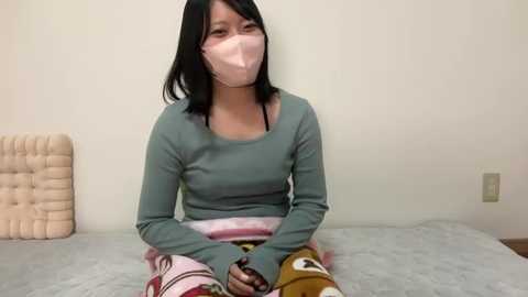 Video of a young Asian woman with long black hair, wearing a teal long-sleeve top, pink mask, and pajamas, sitting on a bed with a beige tufted headboard in a simple, white-walled room.