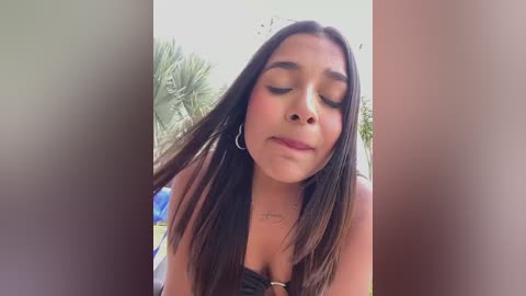 Media: Video of a young woman with medium brown skin and long, straight black hair, wearing a black strapless top and silver hoop earrings, standing outdoors with palm trees in the background.