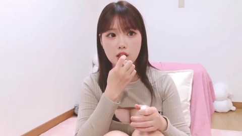 Video of an Asian woman with long black hair, wearing a beige top, eating ice cream in a modern, minimalist bedroom with white walls and a pink bedspread.