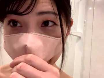 Video of a young Asian woman with fair skin and dark hair, wearing a beige surgical mask, covering her nose and mouth. She appears to be in a bathroom with white tiles and a metal door handle in the background.