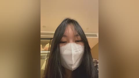 Video of an Asian woman with long black hair, wearing a white surgical mask, standing indoors with beige walls and a metal door in the background.