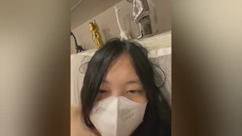 Video of an Asian woman with long black hair, wearing a white face mask, looking directly at the camera. Background includes a white shelf with a golden statue, a silver figurine, and a white statue.