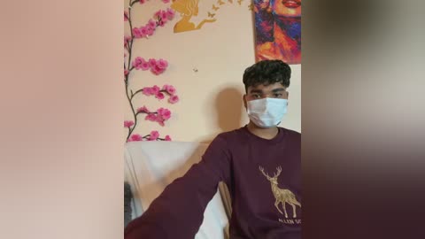 Media: Video of a young South Asian man with short black hair, wearing a maroon sweatshirt with a deer graphic, white mask, and seated in a room with pink floral wallpaper, a colorful painting, and a white chair.