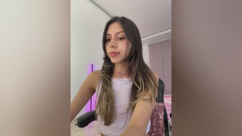 Video of a young Asian woman with long, straight, light brown hair, wearing a white sleeveless top, sitting indoors. Background features a pink-themed room with a gaming chair and a bed, and a purple accent light.
