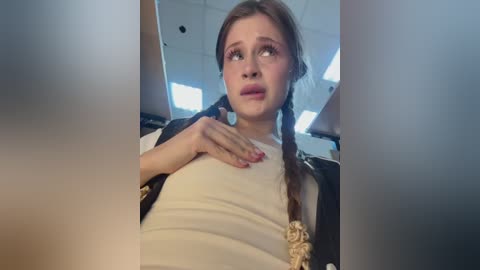 Video of a young Caucasian woman with light skin, brown hair in braids, wearing a white top and black jacket. She has a worried expression, hand on chest, in a modern, brightly lit room with white ceiling tiles.