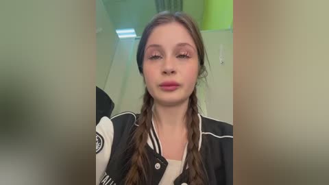 A video of a young woman with long, light brown hair in braids, wearing a black and white varsity jacket, standing indoors with green and yellow walls in the background.