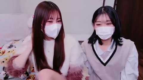Video of two young Asian women in matching white masks and sweaters with fur trim, sitting on a bed with a colorful patterned duvet. They appear to be in a bedroom setting.