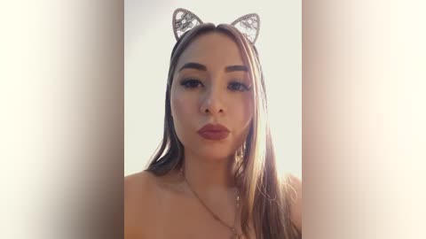 Video of a young Asian woman with long, straight, brown hair, wearing a silver butterfly headband and red lipstick, standing against a bright, soft-focus background.