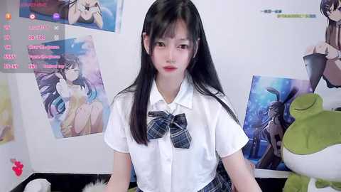 Video of a young East Asian woman with long black hair, wearing a white shirt and plaid bow tie, seated in a room with anime posters and plush toys.