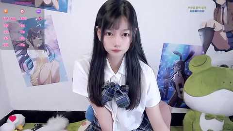 A video of an East Asian young woman with long black hair, wearing a white blouse and blue plaid skirt, seated in a room with anime posters and plush toys.