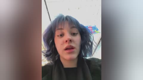 A video of a young woman with shoulder-length, messy, blue hair, wearing a black sweater, taken in a dimly lit, graffiti-covered alleyway.