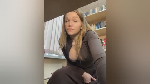 Media: A video of a blonde woman with a medium build, wearing a low-cut black dress, sitting in a room with bookshelves and a window.