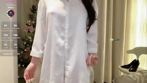 A video of a woman wearing a white, long-sleeved button-up shirt, standing in front of a Christmas tree with ornaments and lights. The background includes beige curtains and a chair with a black shoe on it.
