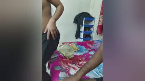 A video shows a topless person in black pants, facing a man in a pink shirt, standing in a cluttered bedroom with a colorful bedspread and a blue shelf with clothes.